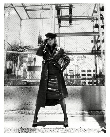 Monochrome fashion shot of a woman wearing a long coat and hat posing outdoors against an industrial background with a fence.