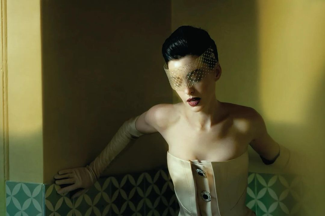 Adult female in strapless top with net veil and long gloves, posing against a patterned wall, in a dimly lit, indoor setting.
