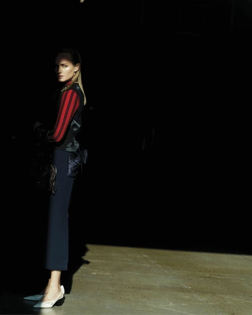 A stylish woman stands in a partially illuminated space, wearing a striped vest, dark trousers, and white heels, looking away from the camera.