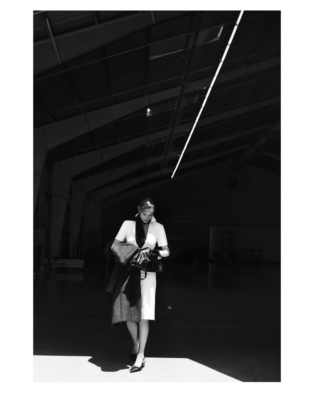 Woman in formal attire walks through an industrial space, wearing a light dress, dark jacket, and heels, in a black and white photograph.