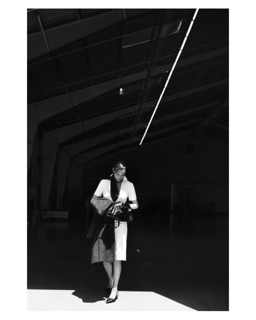 Woman in formal attire walks through an industrial space, wearing a light dress, dark jacket, and heels, in a black and white photograph.