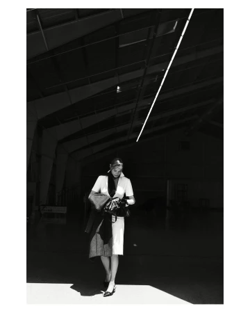 Woman in formal attire walks through an industrial space, wearing a light dress, dark jacket, and heels, in a black and white photograph.