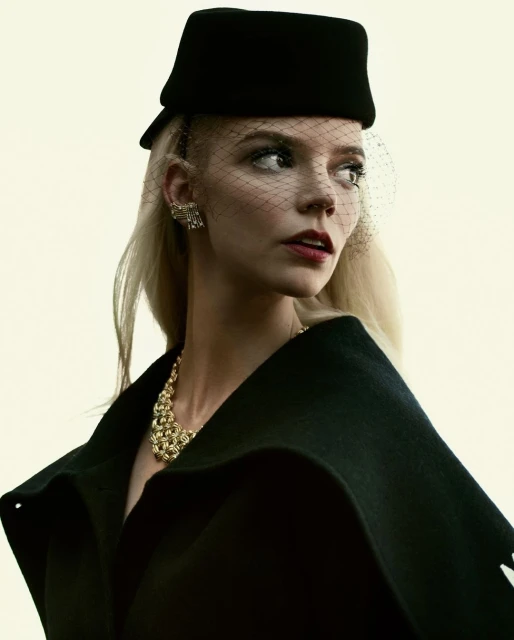Elegant portrait of a blonde woman in a black hat and cape, adorned with a gold necklace and earrings, set against a soft white background.