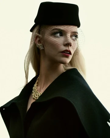 Elegant portrait of a blonde woman in a black hat and cape, adorned with a gold necklace and earrings, set against a soft white background.