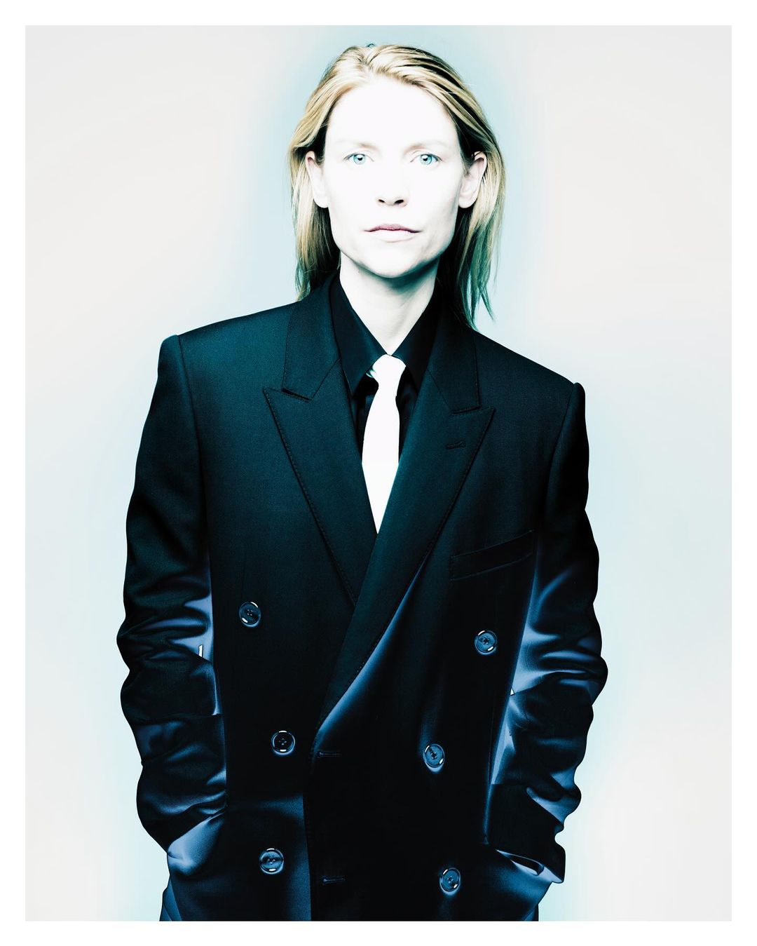 Portrait of Claire Danes wearing a dark double-breasted suit jacket with a white tie against a pale background.