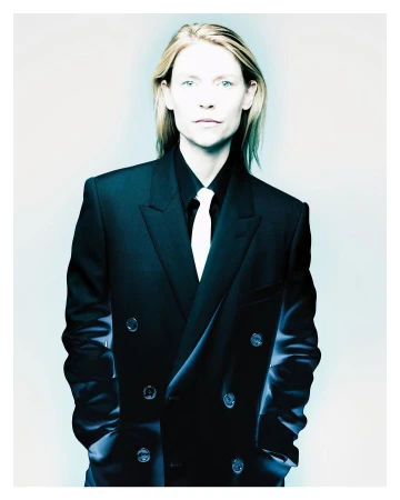 Portrait of Claire Danes wearing a dark double-breasted suit jacket with a white tie against a pale background.