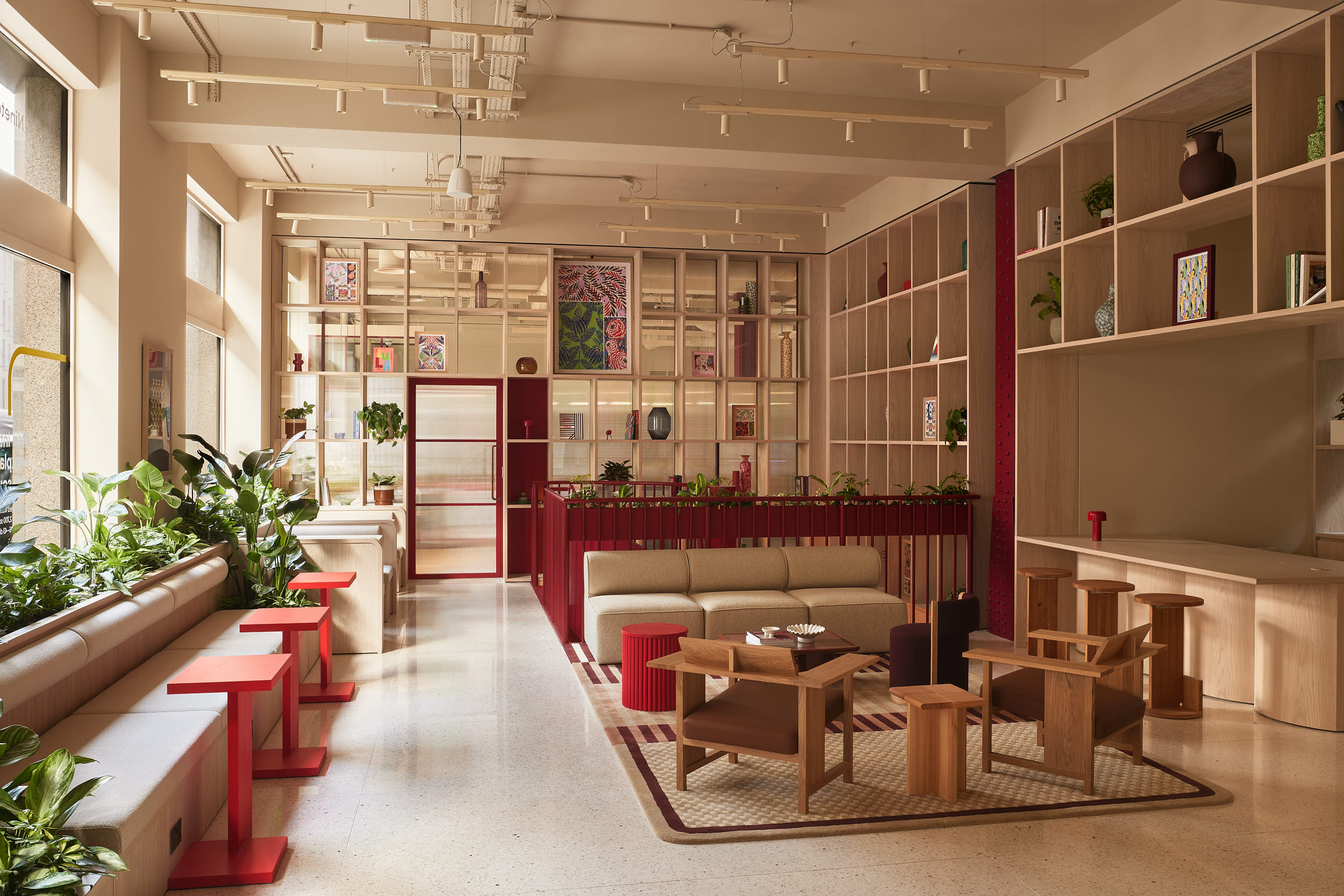 Modern open-plan office lounge interior with light wood shelving, neutral sofa, wooden armchairs, and red accent side tables.