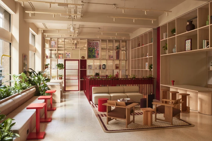 Modern open-plan office lounge interior with light wood shelving, neutral sofa, wooden armchairs, and red accent side tables.
