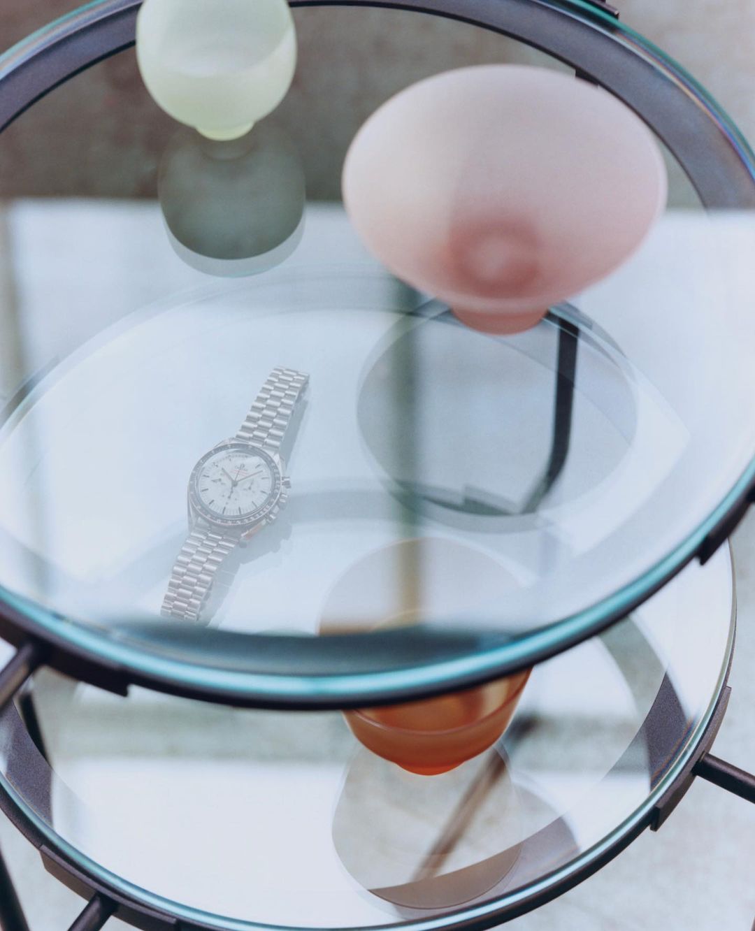 Wristwatch with a metal band on a tiered glass table next to decorative spheres in shades of green, white, and peach. Clean product shot.