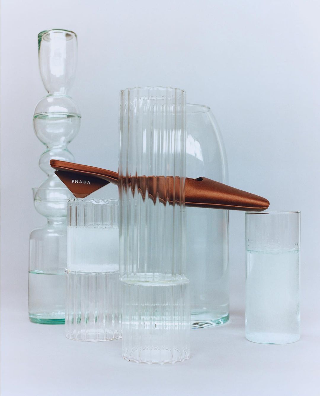 Tan Prada mule resting atop clear glass vases filled with water against a soft white backdrop in a minimalist still life composition.