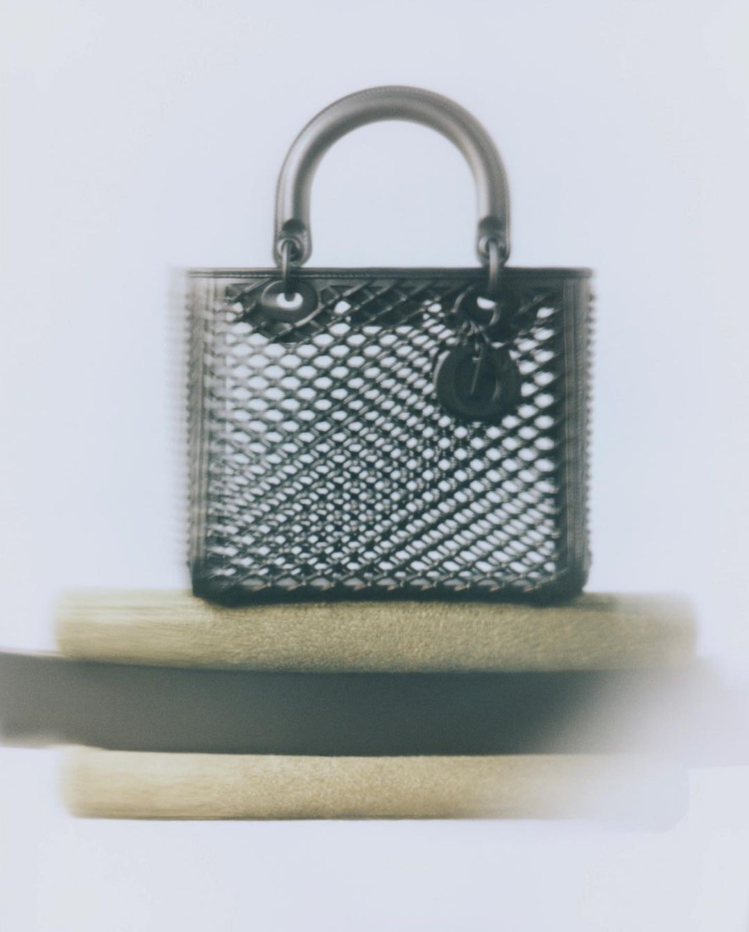 Close-up studio shot of a metallic mesh Lady Dior handbag with silver hardware, resting on blurry brown cushions against white.