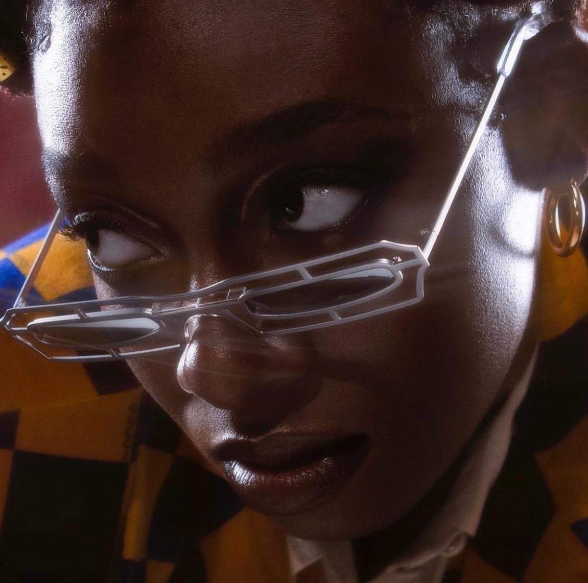 Close-up portrait of a stylish young woman with dark skin, wearing modern glasses and gold hoop earrings, in a dimly lit studio.