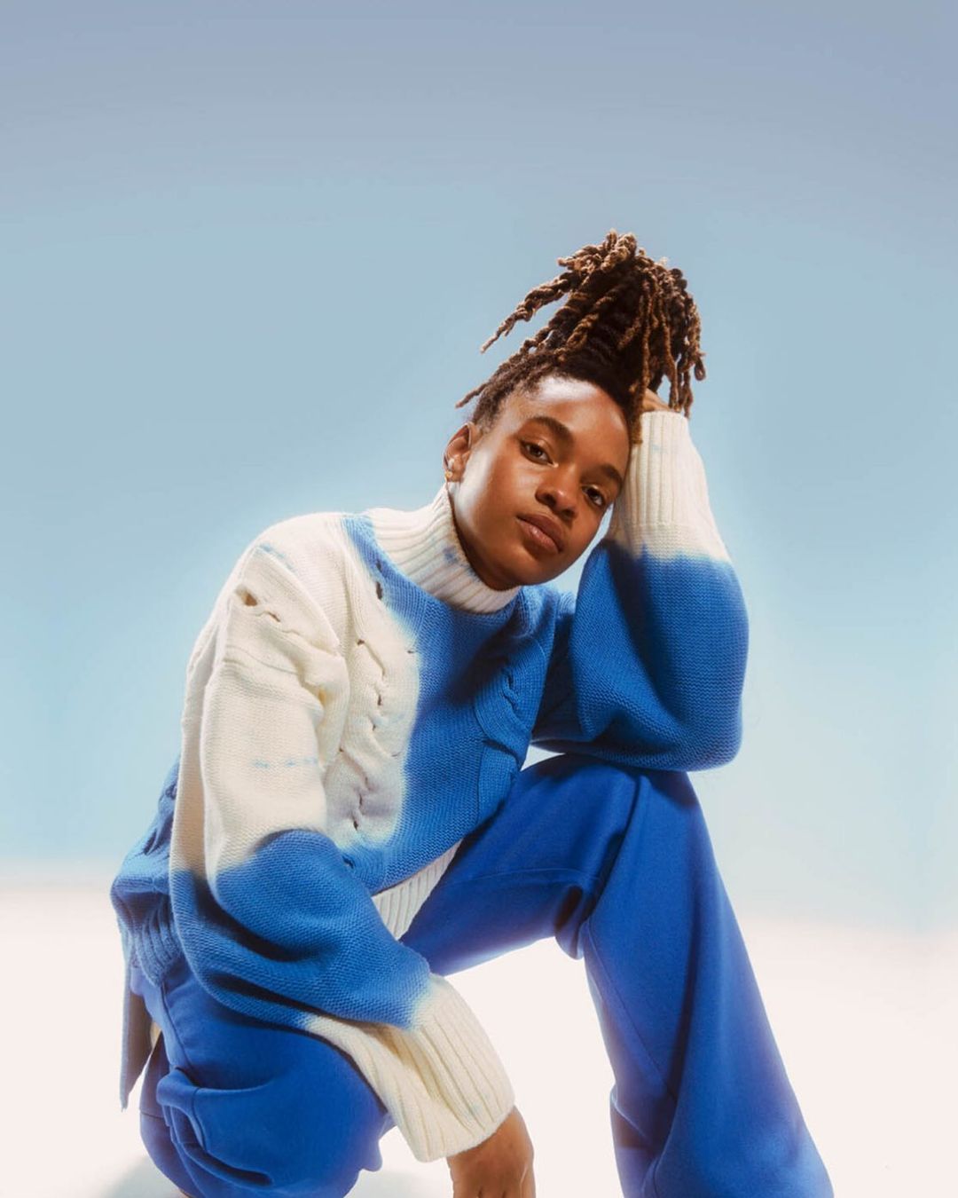 A woman with dreadlocks, wearing a blue and white sweater and blue pants, poses in a studio with a light blue and white gradient background.