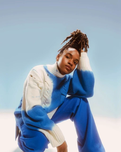 A woman with dreadlocks, wearing a blue and white sweater and blue pants, poses in a studio with a light blue and white gradient background.