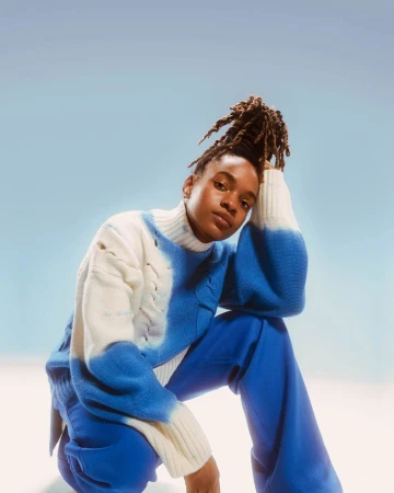 A woman with dreadlocks, wearing a blue and white sweater and blue pants, poses in a studio with a light blue and white gradient background.