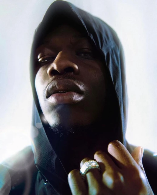 Portrait of an African-American man wearing a hooded sweatshirt with a large diamond ring, photographed at eye-level with a white background.