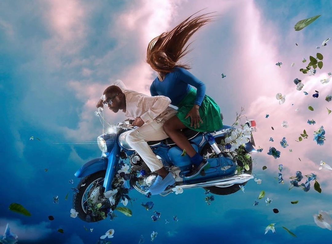 Man and woman riding a vintage blue motorcycle through a swirling sky filled with scattered flowers and clouds, captured by Nwaka Okparaeke.