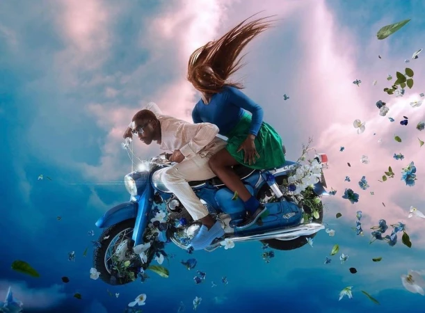 Man and woman riding a vintage blue motorcycle through a swirling sky filled with scattered flowers and clouds, captured by Nwaka Okparaeke.