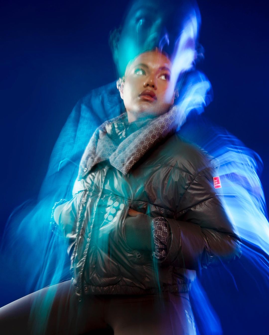 A young woman with short hair poses in a shiny black puffer jacket against a blue background with motion blur effects, looking upward.