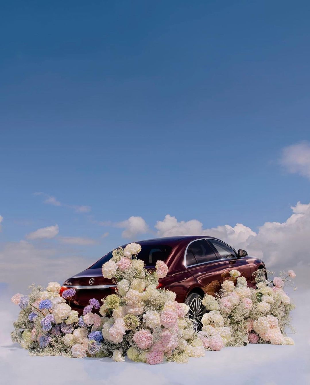 A burgundy car surrounded by pastel flowers against a blue sky with clouds. Automotive photography. Artistic car presentation.