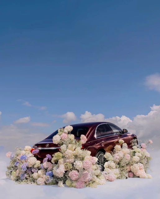A burgundy car surrounded by pastel flowers against a blue sky with clouds. Automotive photography. Artistic car presentation.