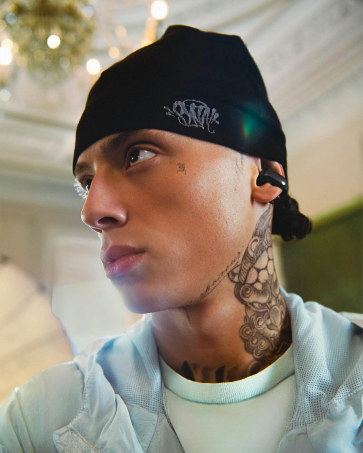 Portrait of a young adult male wearing a black beanie, light blue jacket, and wireless earbuds, showcasing neck tattoo in an indoor setting.