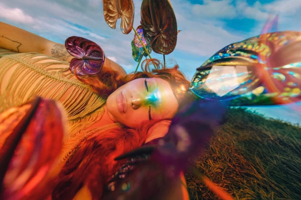 A woman lies on the ground with colorful makeup and flowers, creating a surreal and vibrant outdoor portrait with grass and sky visible.