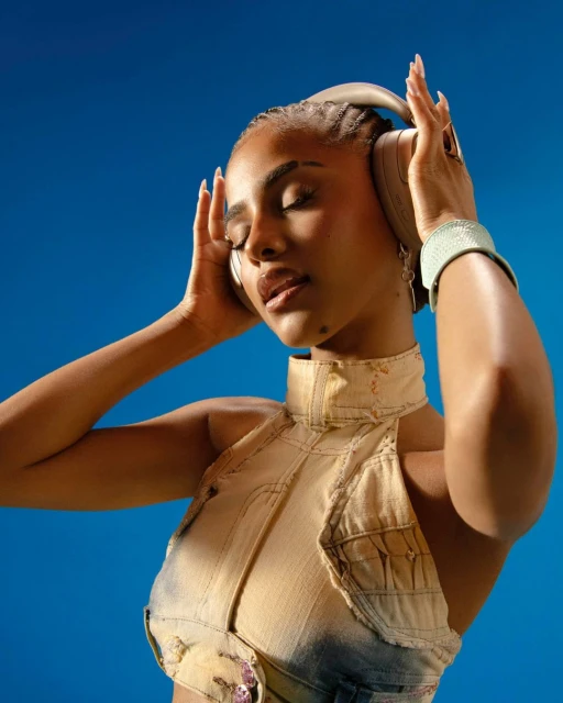 Portrait of a young woman wearing headphones and a denim top, eyes closed, against a blue backdrop in a music or fashion context.