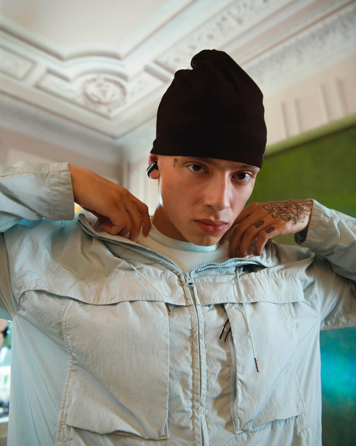 Portrait of a young adult man wearing a black beanie and a light blue jacket indoors with decorative ceiling, posing for the camera.