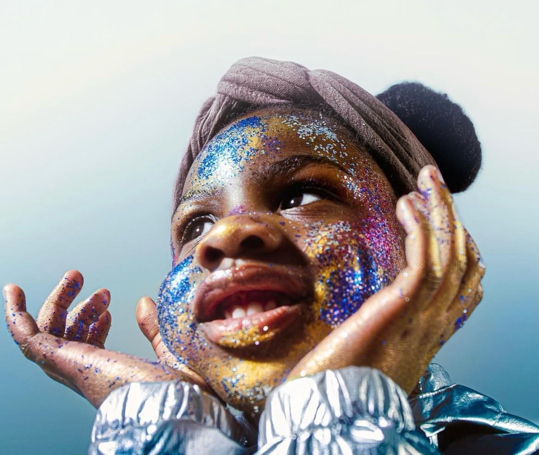 Young girl with sparkly glitter on face and hands, wearing a headwrap and silvery jacket, looking upward in a softly lit studio.