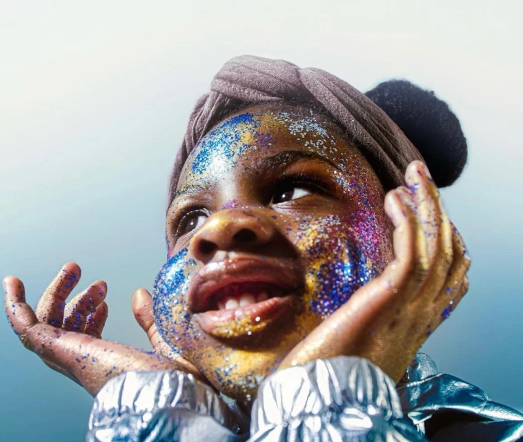 Young girl with sparkly glitter on face and hands, wearing a headwrap and silvery jacket, looking upward in a softly lit studio.