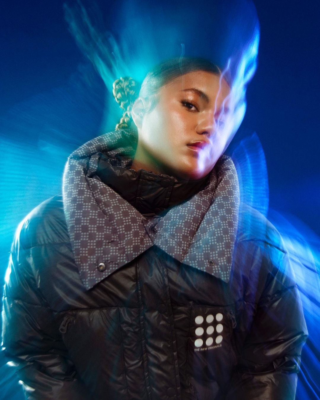 A cool portrait of a woman in a black puffer jacket with braids, set against a vibrant blue background with dynamic lighting effects.