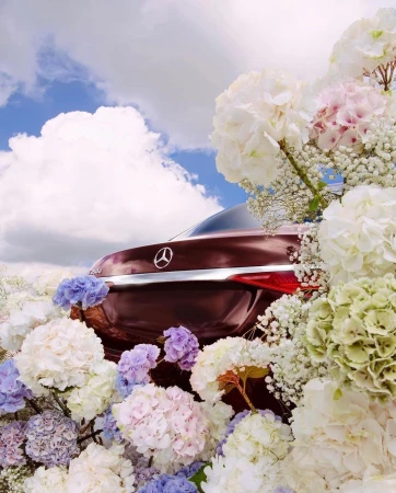 Rear view of a burgundy car surrounded by white, pink, and purple hydrangeas against a cloudy blue sky, creating a vibrant floral automotive scene.