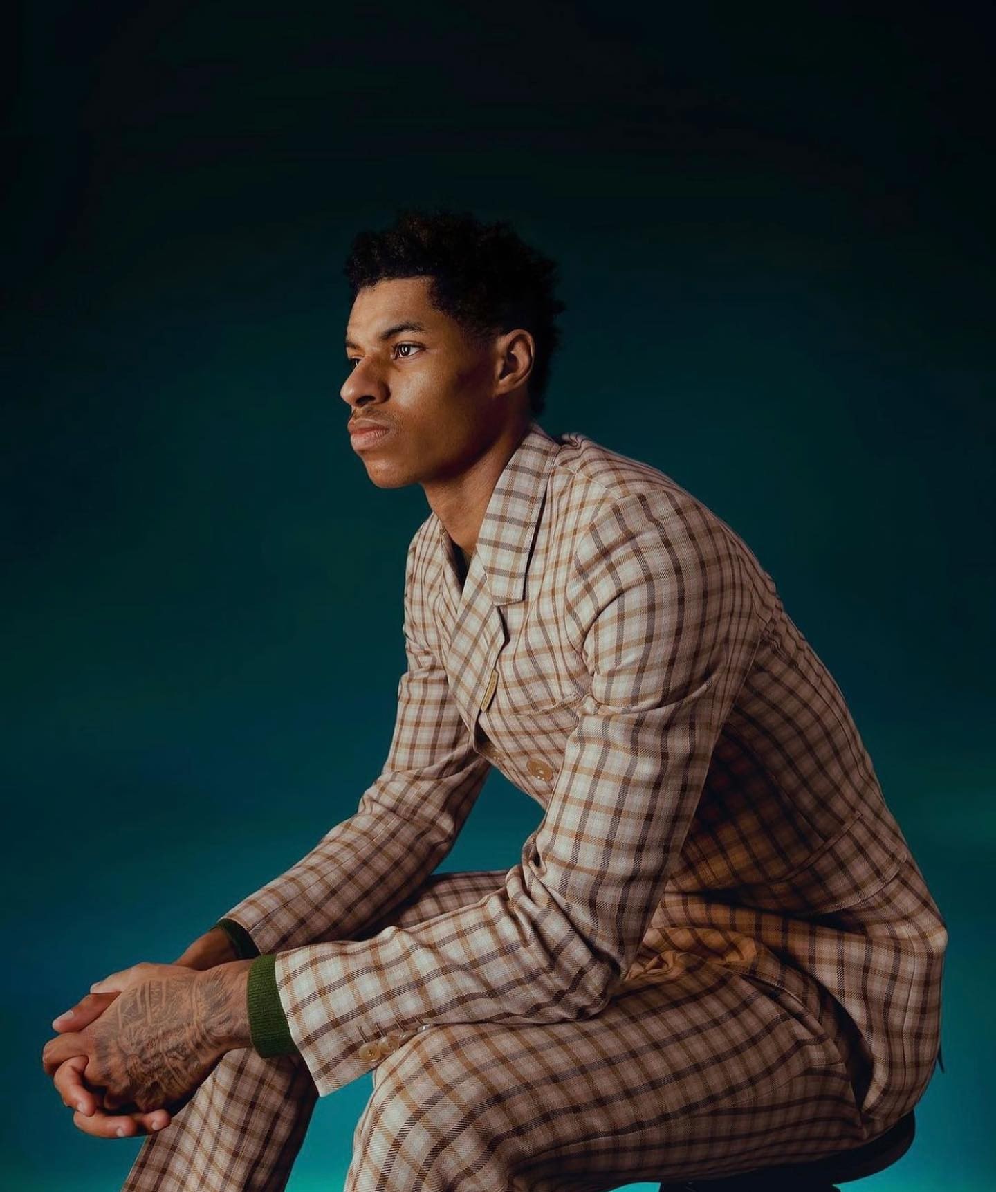 Portrait of a serious adult male in a brown plaid suit sitting in a photo studio. Close-up side view against a teal background.