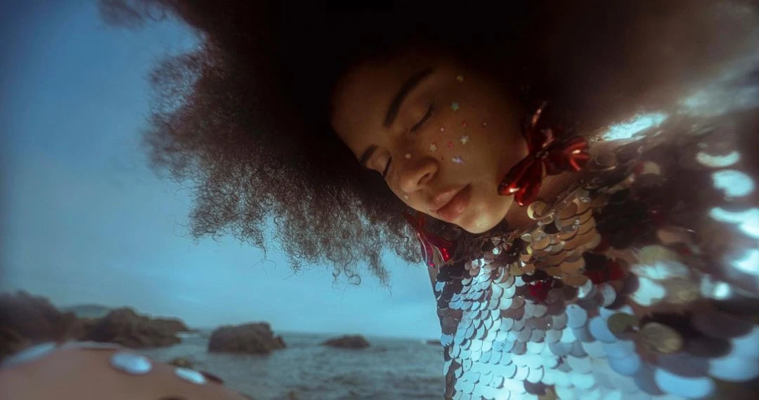Close up shot of an adult woman with glittery face makeup wearing a shiny, sequined top with large afro, standing near the ocean.