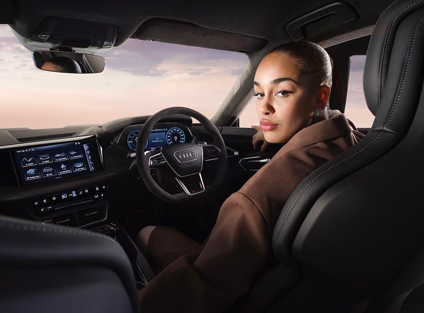 Adult female inside an Audi e-tron GT, showcasing luxury car interior, dashboard, and steering wheel with a view of a pink sky.