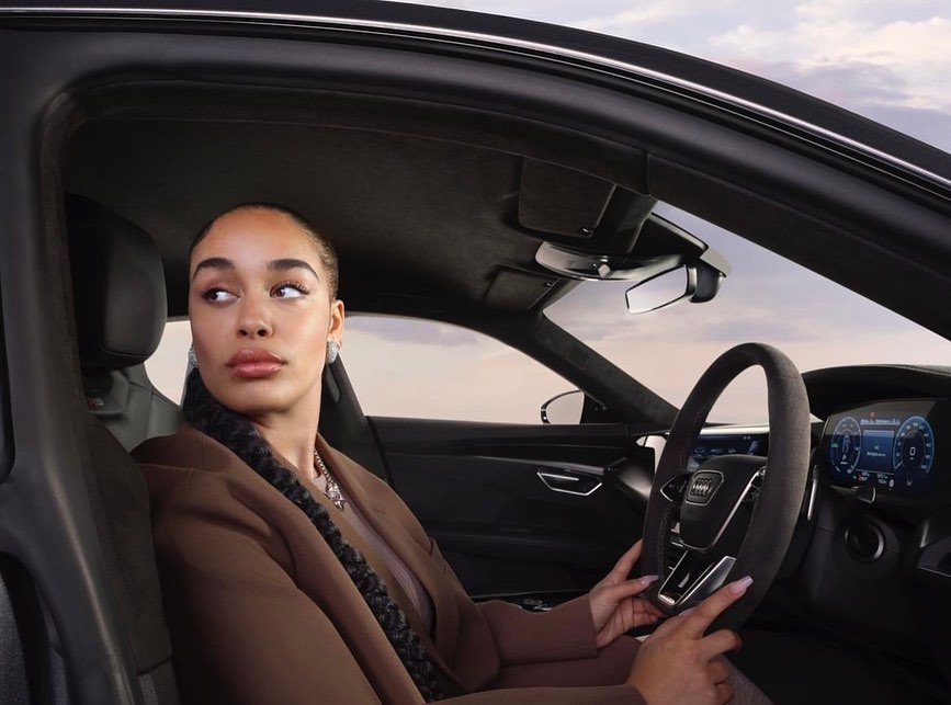 An adult female is sitting in the driver's seat of an Audi, looking out of the car window with a neutral expression, in an automotive setting.
