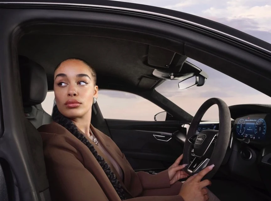 An adult female is sitting in the driver's seat of an Audi, looking out of the car window with a neutral expression, in an automotive setting.