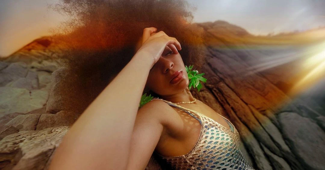 Woman with afro hairstyle lies on rocky surface near water wearing a shimmering top with lens flare and mountains in the background.