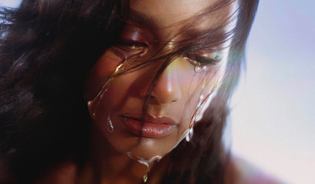Close-up portrait of a woman with dark hair and shimmering makeup, featuring water droplets cascading down her face in an atmospheric style.