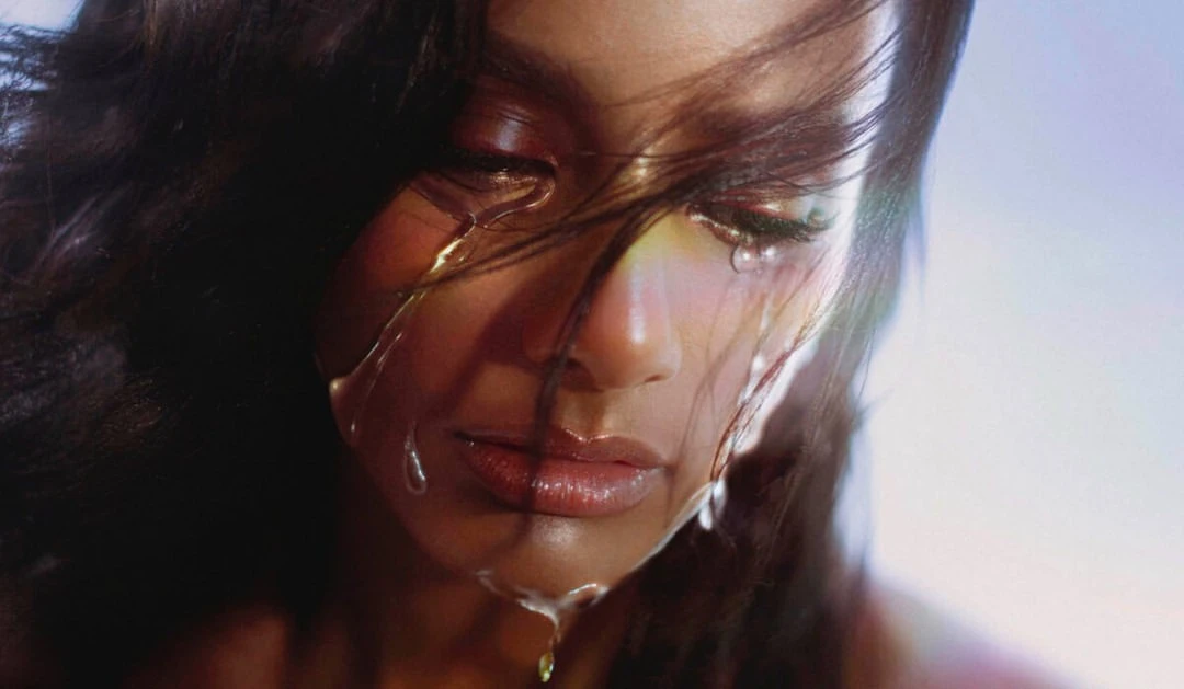 Close-up portrait of a woman with dark hair and shimmering makeup, featuring water droplets cascading down her face in an atmospheric style.