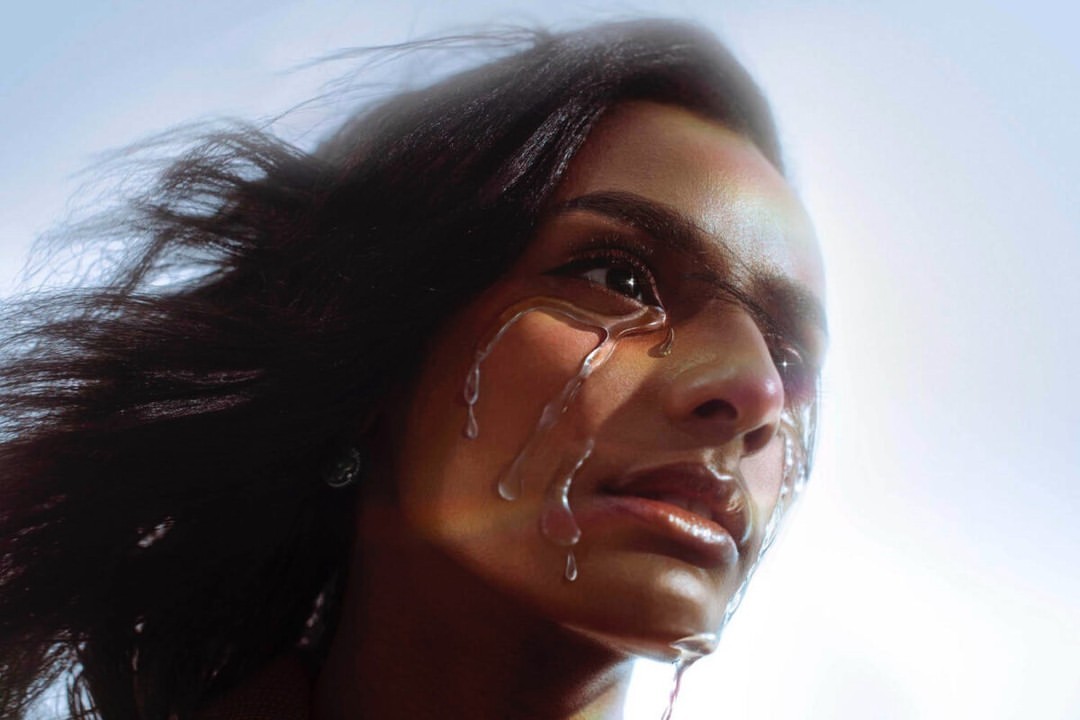 Close-up portrait of a woman with liquid dripping down her face, creating a glossy, textured effect against a bright sky background.
