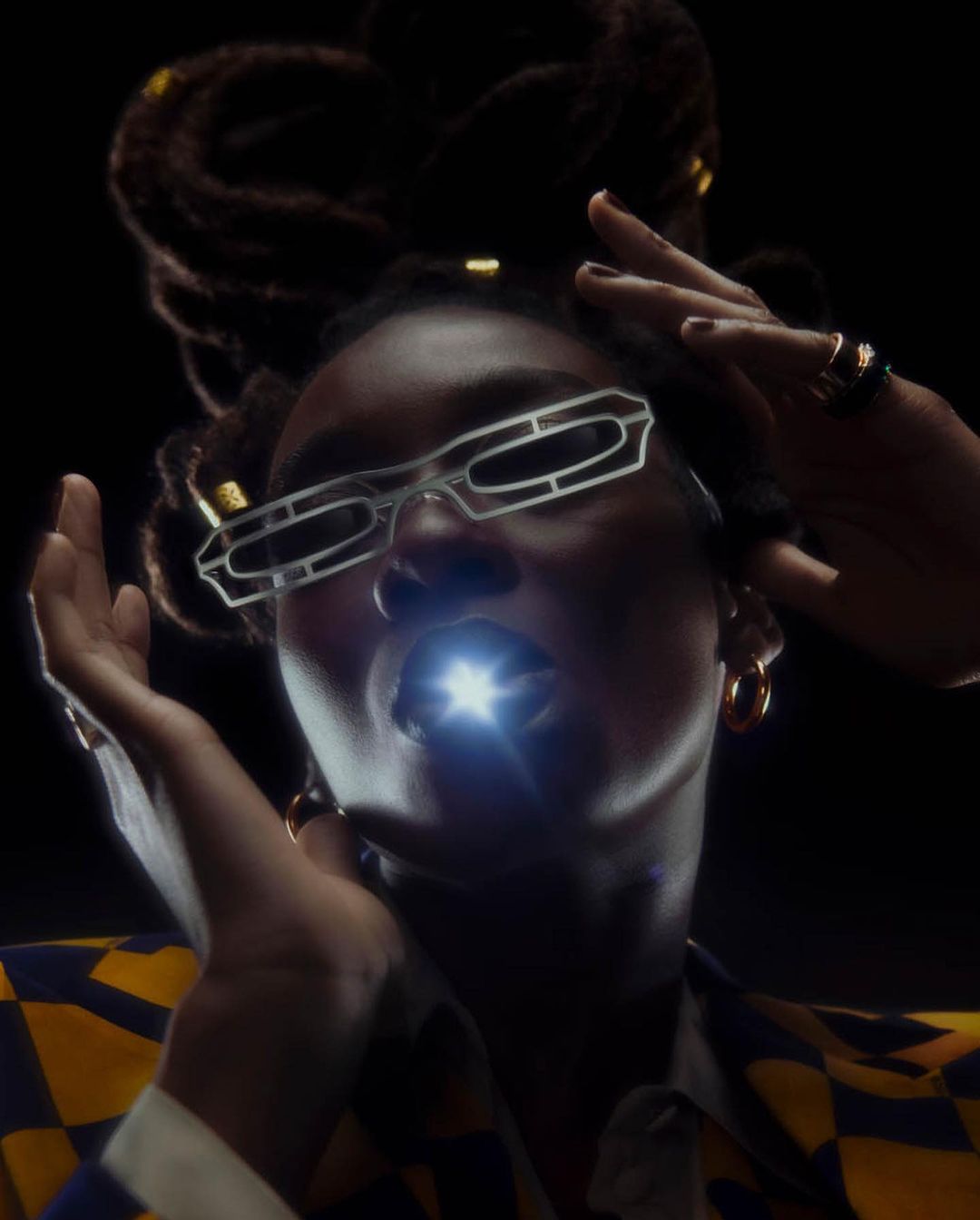 Close-up portrait of a Black woman with glasses, dreadlocks, a blue and yellow jacket, and a light shining from inside her mouth.