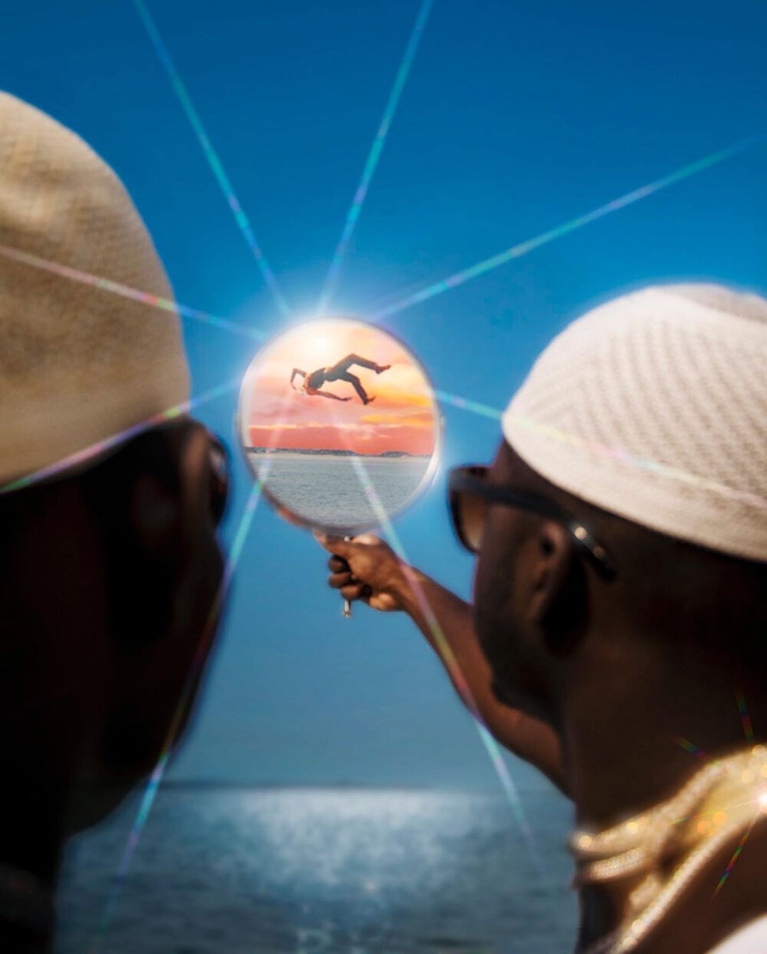 Two men look at a hand mirror reflecting a falling figure against a sunset ocean background on a clear day with beams of light.