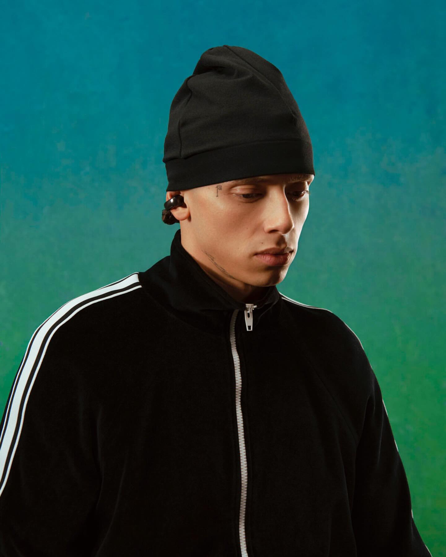 Portrait of a young adult male wearing a black beanie and track jacket against a gradient blue and green background, studio lighting.