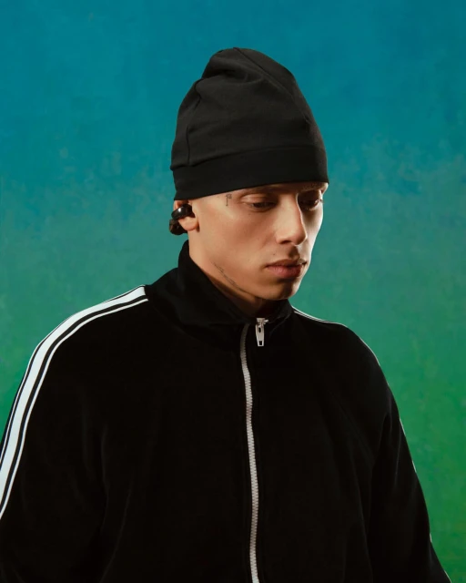 Portrait of a young adult male wearing a black beanie and track jacket against a gradient blue and green background, studio lighting.