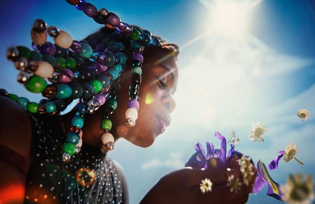 Adult female smelling flowers in outdoor close-up, sunlight, blue sky background. Braids with beads and sparkling top.