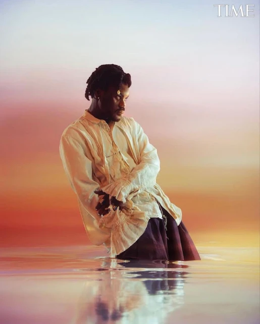 Portrait of an African American man waist deep in shallow water against a gradient sunset backdrop, showcasing editorial fashion.