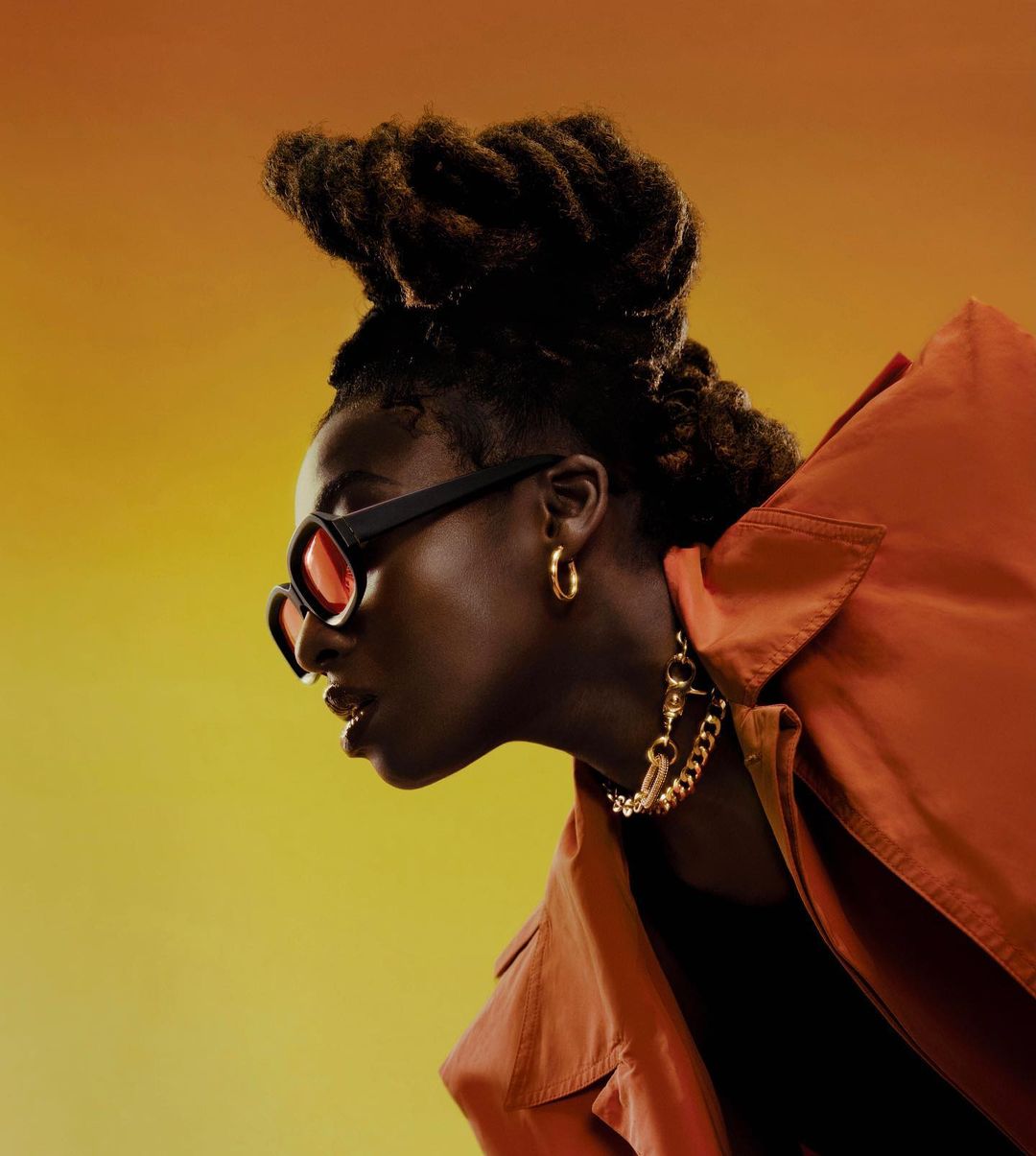 Profile shot of a Black adult female with locs, sunglasses, a rust-colored jacket, and gold jewelry against an orange gradient background.