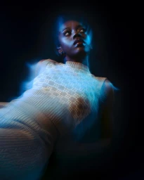 Portrait of a Black woman wearing a patterned sleeveless knit top with bright blue light tracing her form against a dark background